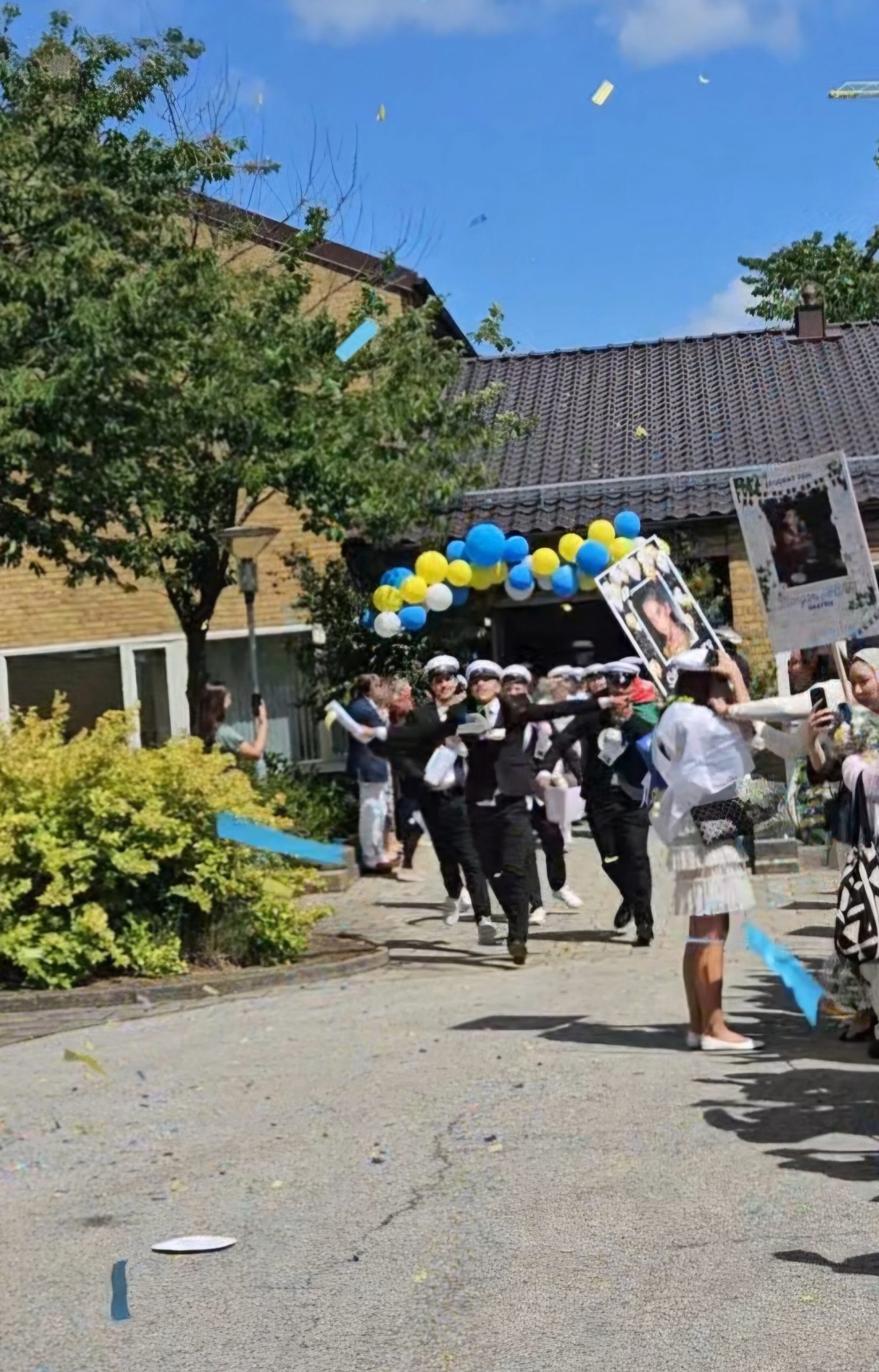 High school students in white caps run out of their school, greeted by cheering family and friends holding signs and balloons.