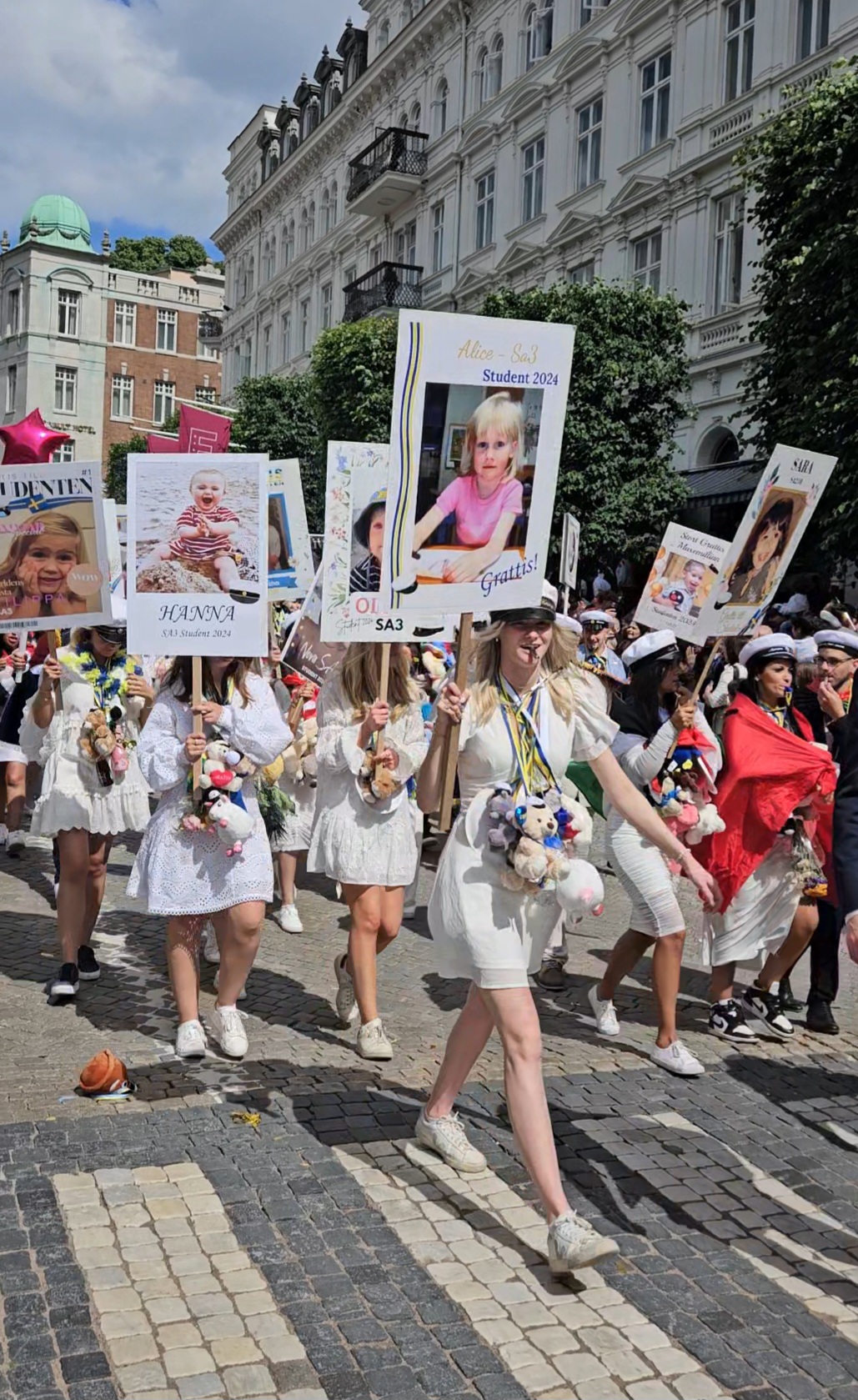 "Girls in white dresses walk through a festive crowd-lined street, celebrating their graduation day in the city center