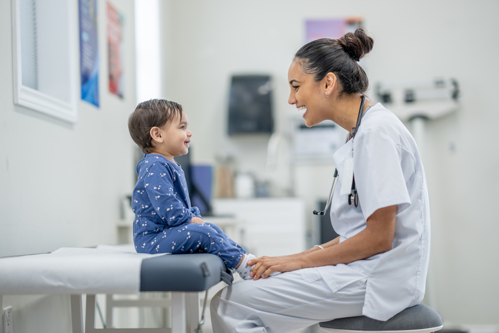 A boy toddler sitting on an examination table while a female doctor checks him with a stethoscope. The child appears calm and attentive, and the doctor is smiling warmly, creating a reassuring atmosphere