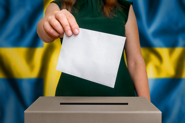 A person placing their ballot into a voting box with the Swedish flag in the background